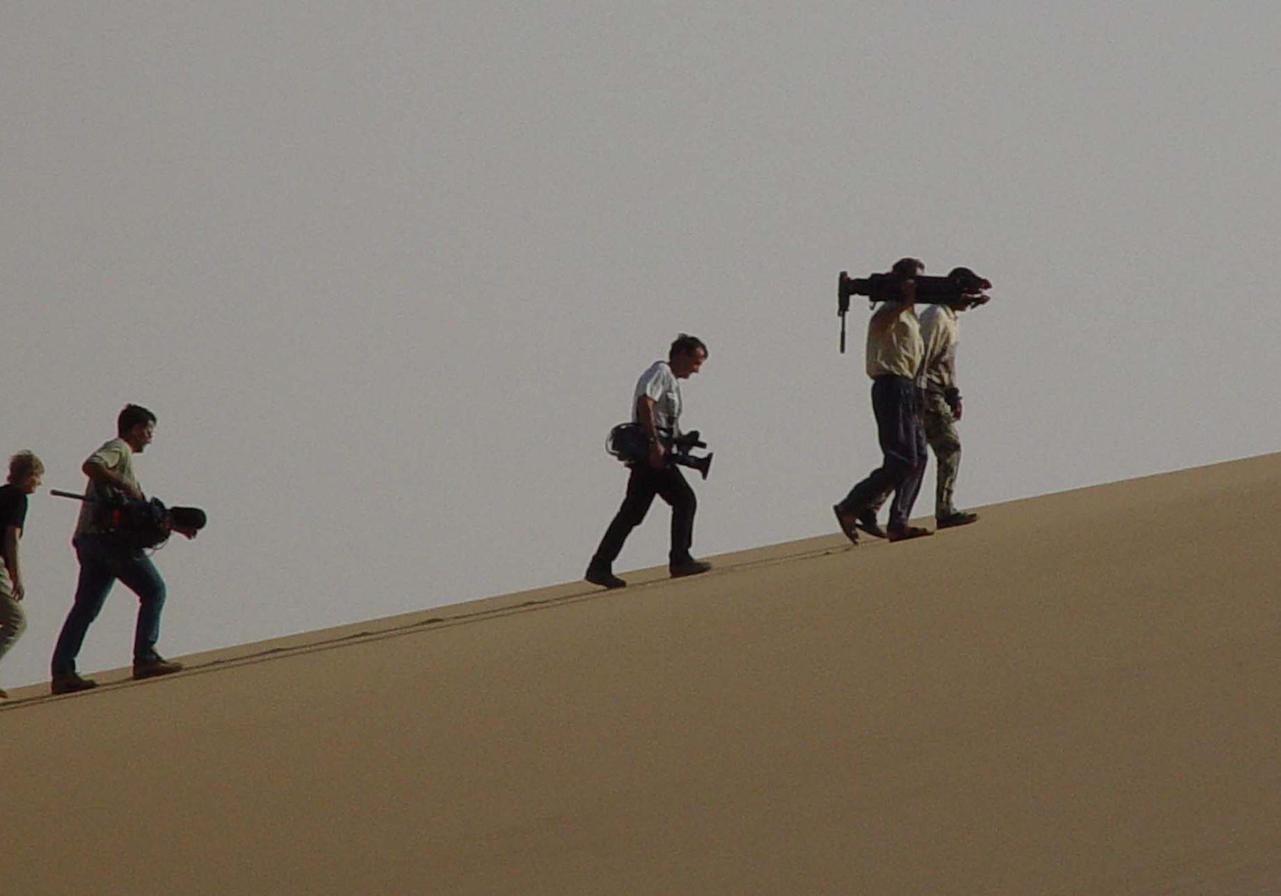 Film crew carrying equipment over sand dune